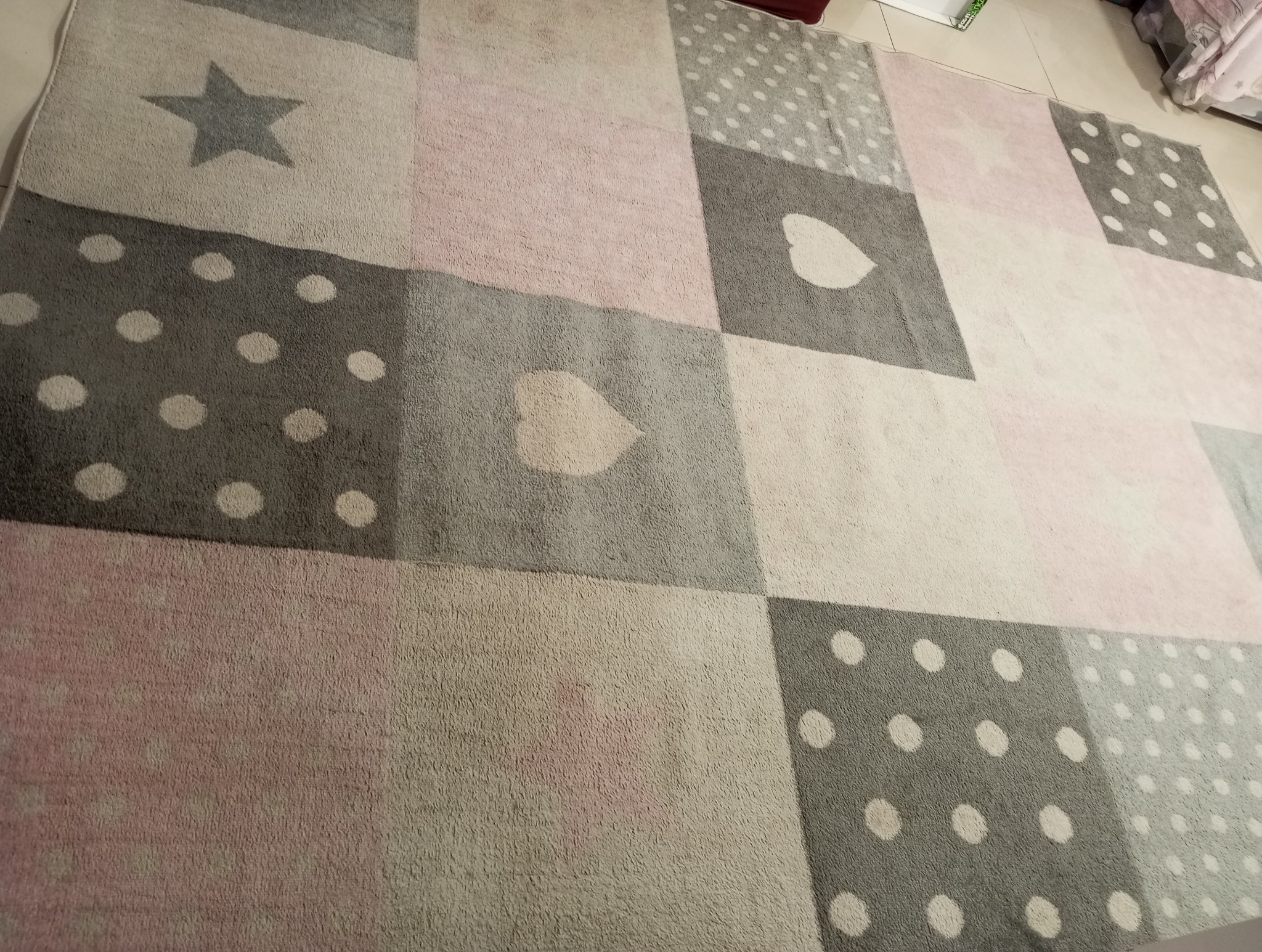 Dziecięcy dywan w pastelowych odcieniach szarości i różu, wzór w kwadraty z gwiazdkami, serduszkami i groszkami, widoczne ślady użytkowania.