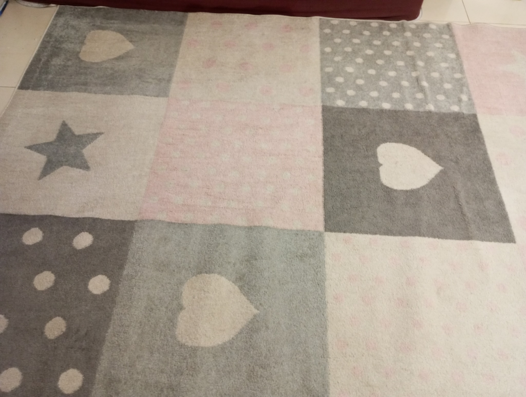 Fragment wzorzystego dywanu w odcieniach szarości, różu i bieli z motywami serc, gwiazd i kropek, leżącego na podłodze.