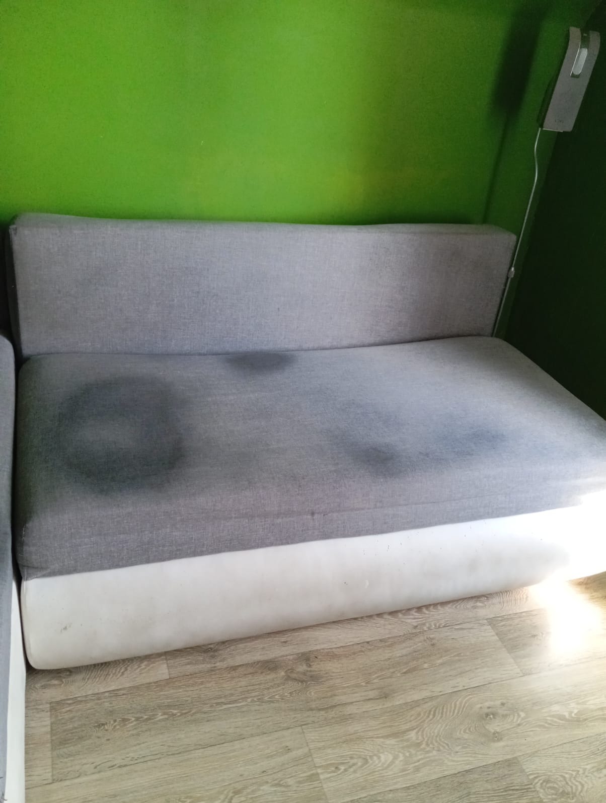 Szara kanapa z wyraźnymi plamami na siedzisku, biały dół, na tle zielonej ściany i drewnianej podłogi.