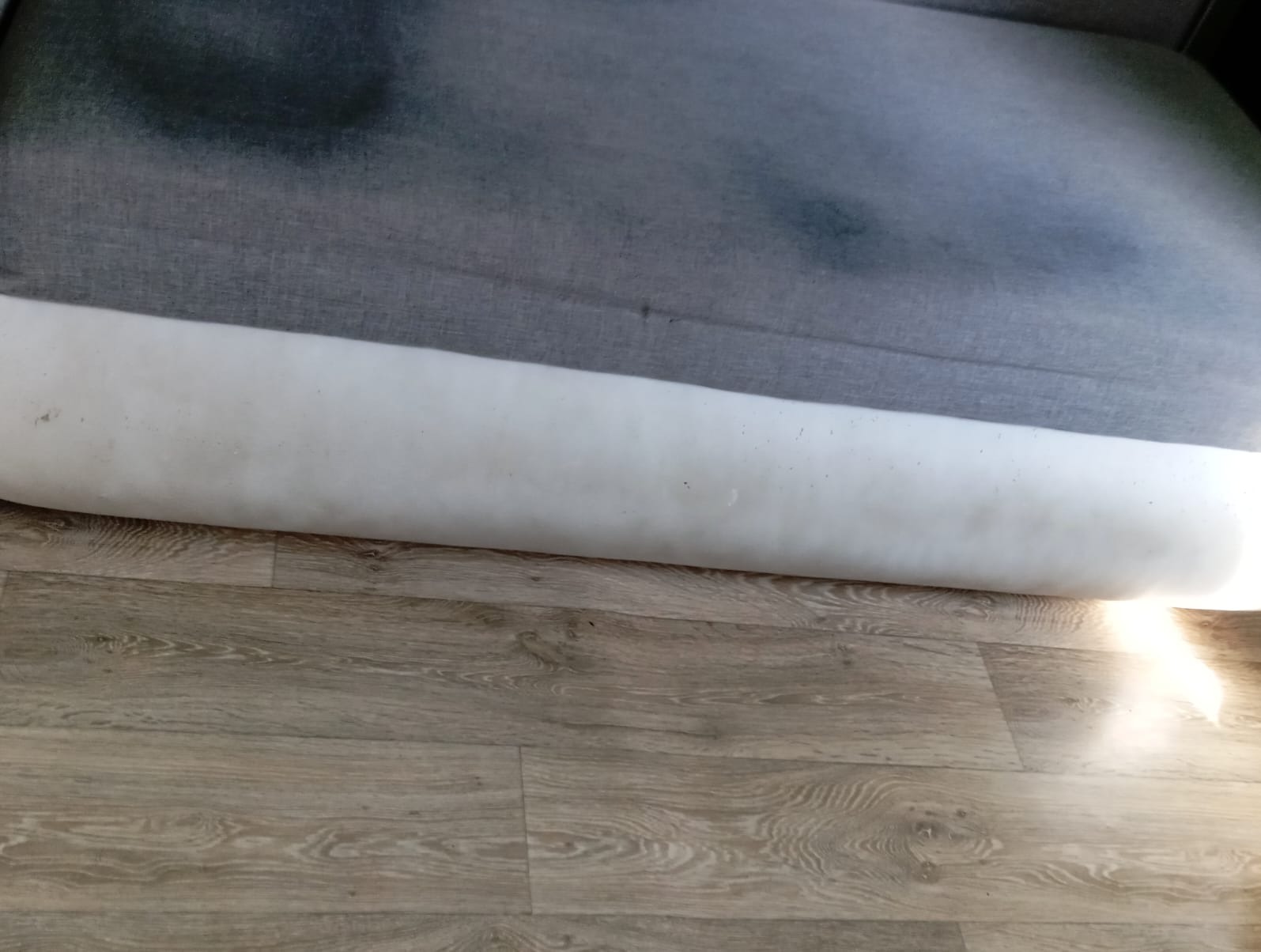 Szary narożnik z widocznymi plamami na siedzisku i zabrudzonym białym bokiem, ustawiony na panelach podłogowych.