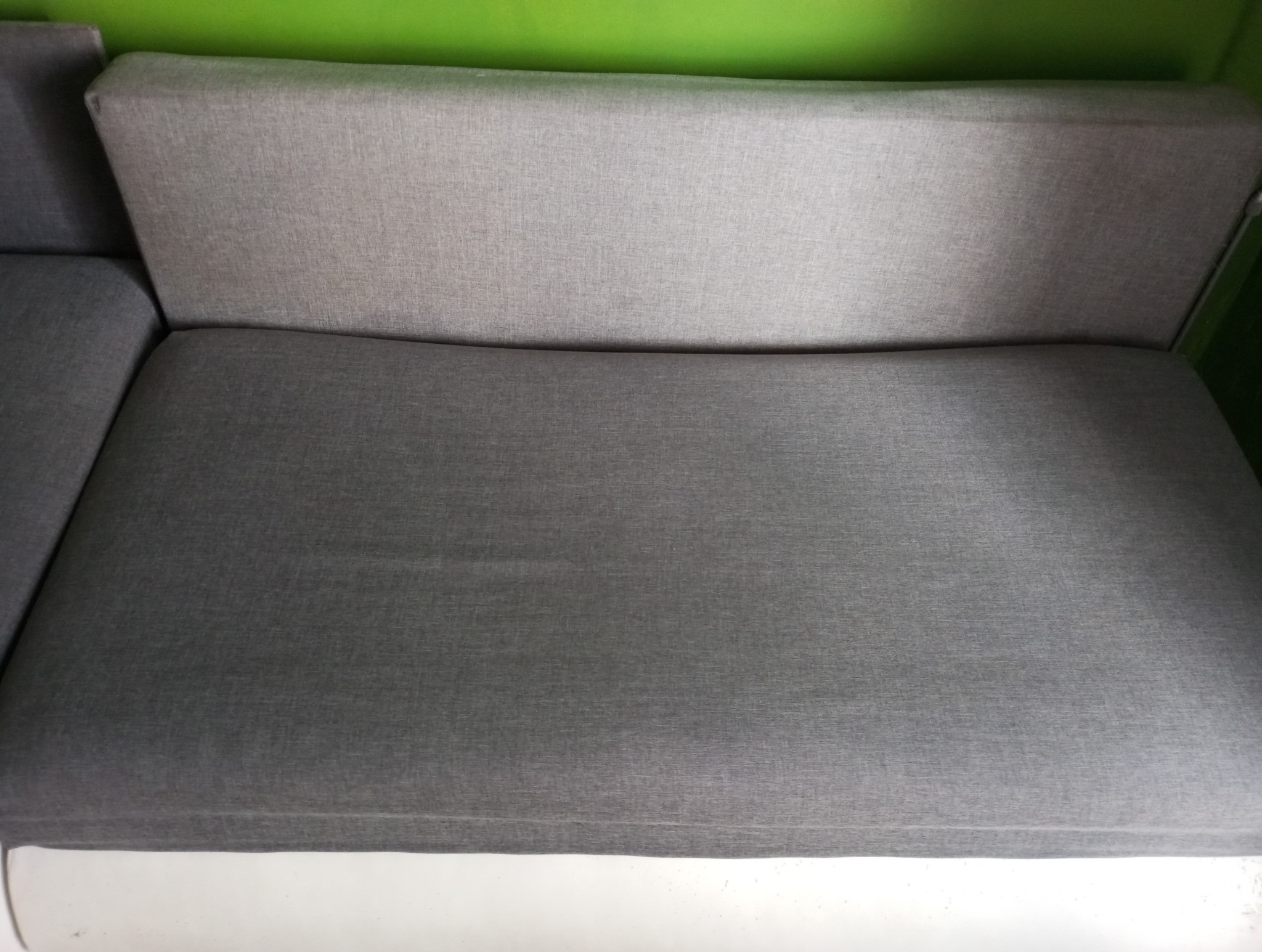 Szara, tekstylna kanapa z widocznym oparciem i siedziskiem, ustawiona na białej podłodze na tle zielonej ściany.