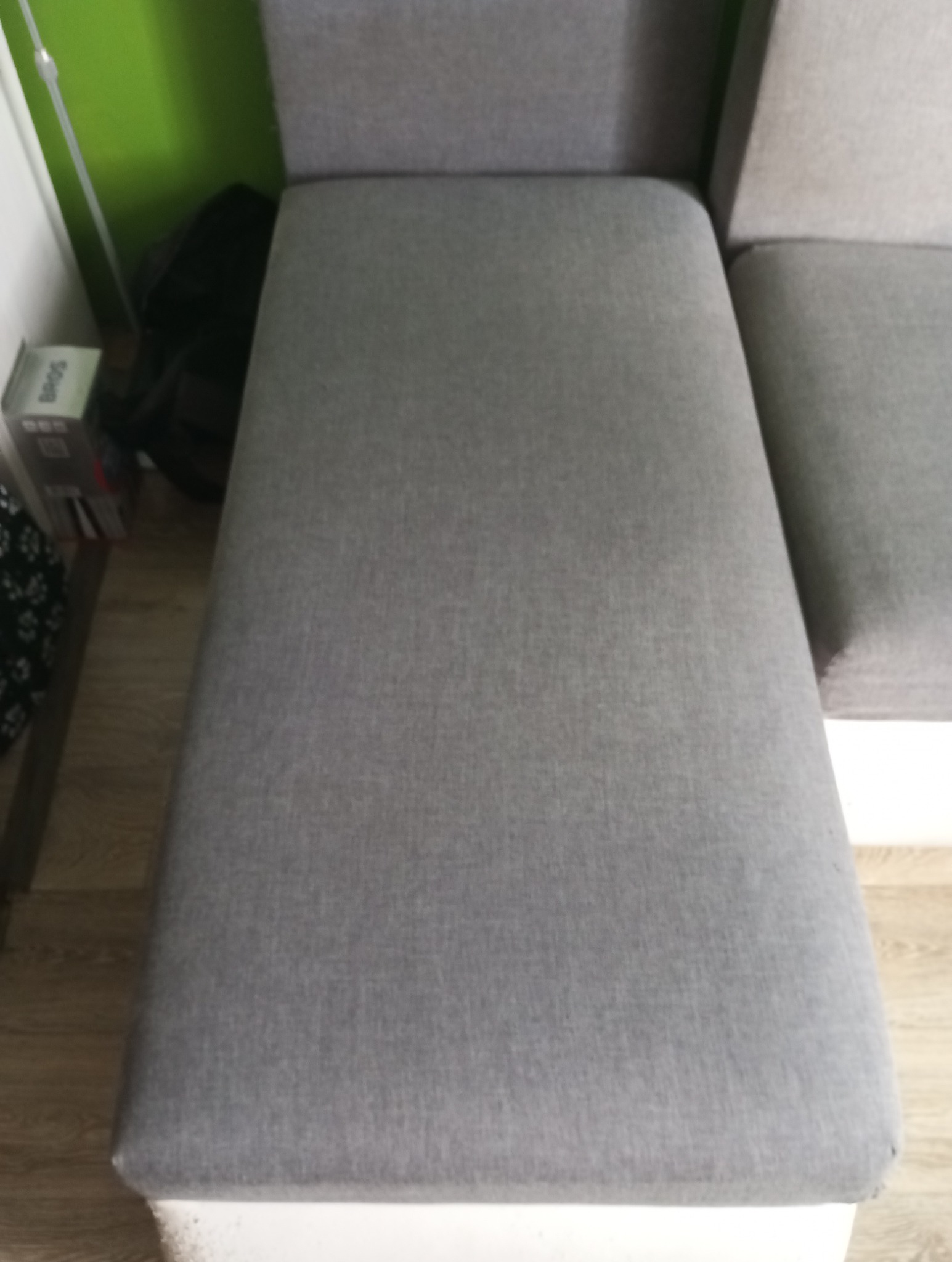 Fragment szarej kanapy modułowej z widoczną fakturą tkaniny i białym obramowaniem, obok ściana w odcieniach zieleni oraz kartonowe pudełko na podłodze.