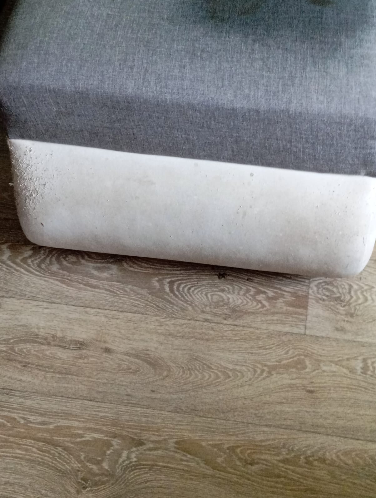Szary narożnik z jasną, zabrudzoną podstawą, stojący na panelach podłogowych z wyraźnym wzorem drewna.