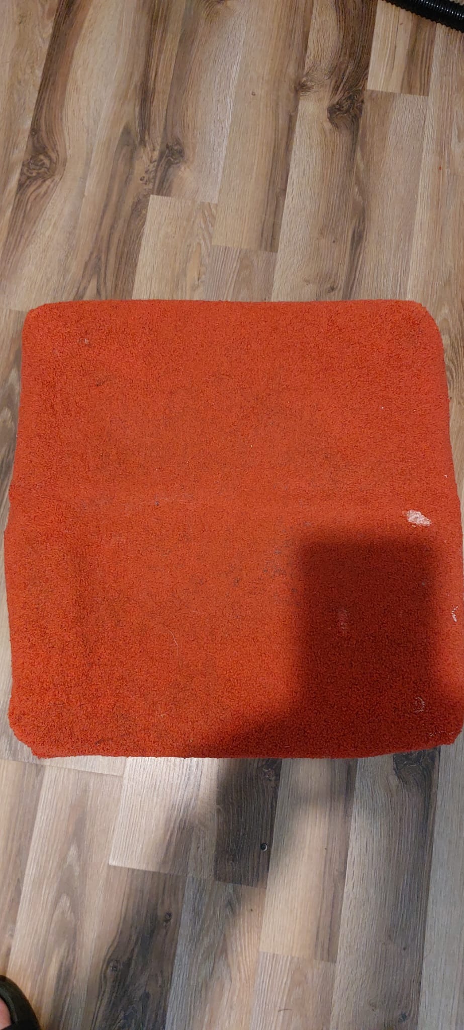 Pomarańczowy, prostokątny dywanik z krótkim włosiem, leżący na panelach podłogowych o wzorze drewna, widoczne plamy i ślady użytkowania.