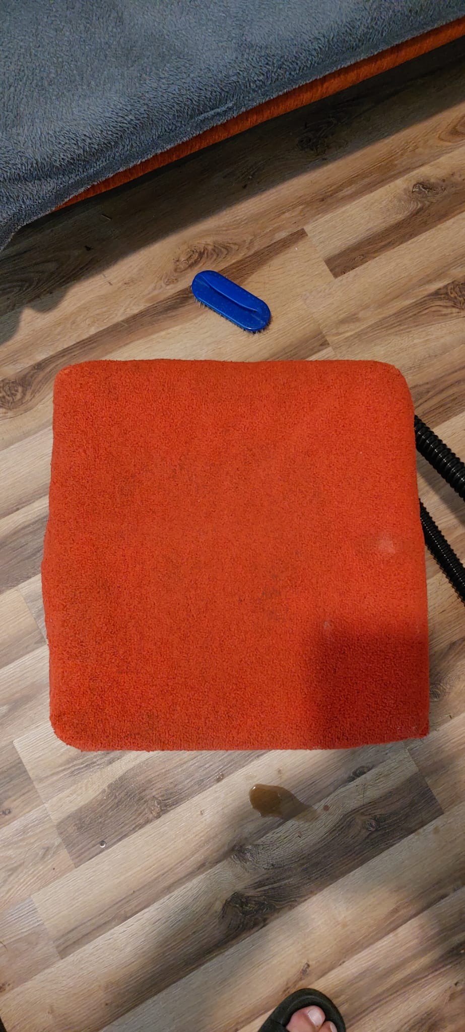 Czerwony, kwadratowy puf z widocznymi plamami i szczotką do czyszczenia na podłodze imitującej drewno, obok czarnego węża odkurzacza.