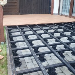 Tarasy Andrii Pohlod - Montaż podkonstrukcji tarasu na regulowanych wspornikach. Częściowo ułożone deski, widoczne legary i kostka brukowa. Na trawie stos desek i karton.