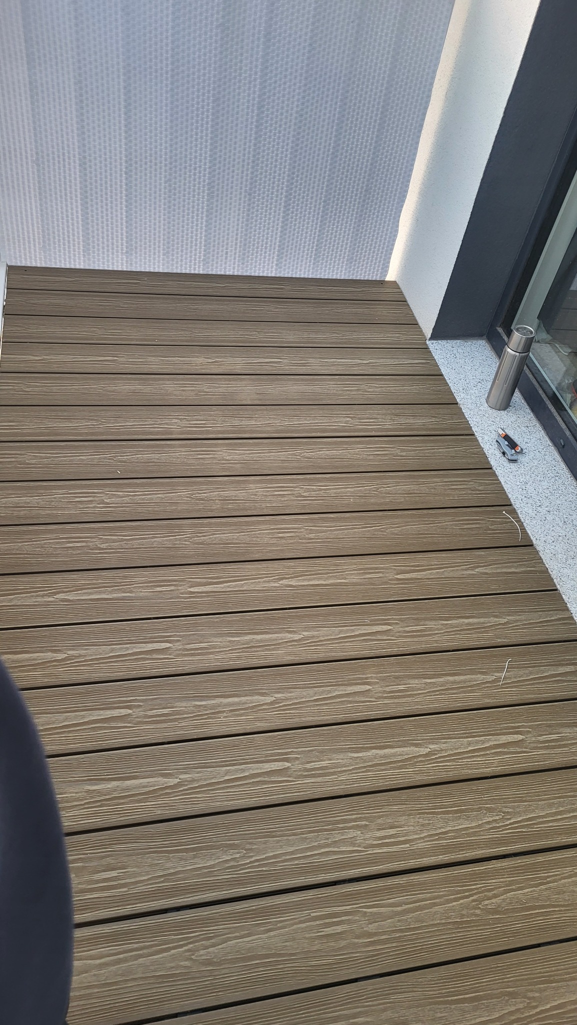 Nowoczesny taras z desek w odcieniach brązu, widok z góry. Na obrzeżach widoczny fragment okna i termosu, co dodaje realizmu i sugeruje użytkowanie.