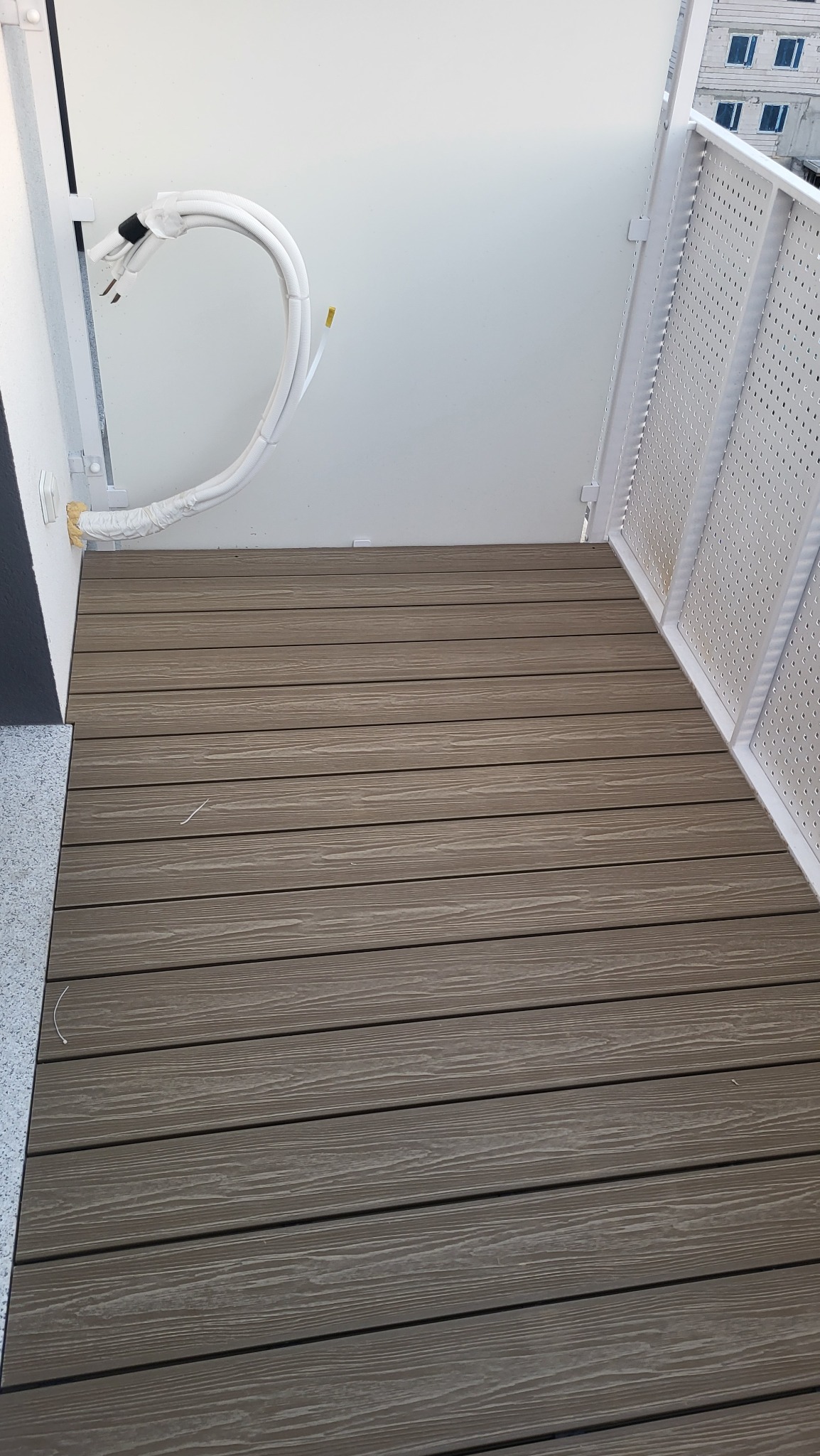 Balkon z brązową podłogą z desek, białą balustradą i widocznymi przewodami klimatyzacji przy ścianie. Ujęcie z góry, podkreślające detale wykończenia.