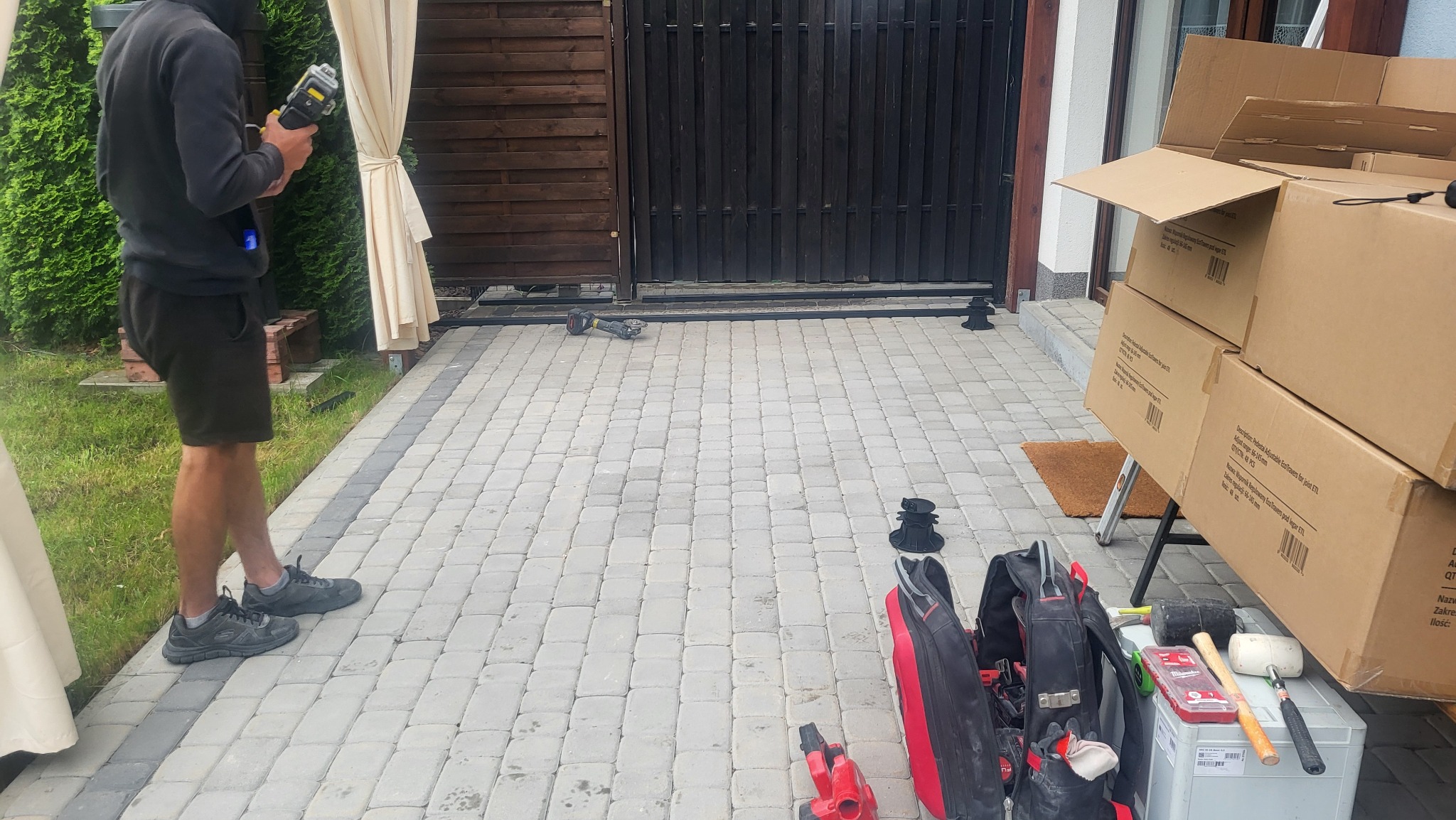 Montaż tarasu z kostki brukowej: pracownik z narzędziem, widoczne elementy konstrukcyjne, skrzynki z narzędziami i materiałami. Etap budowy, kostka ułożona.