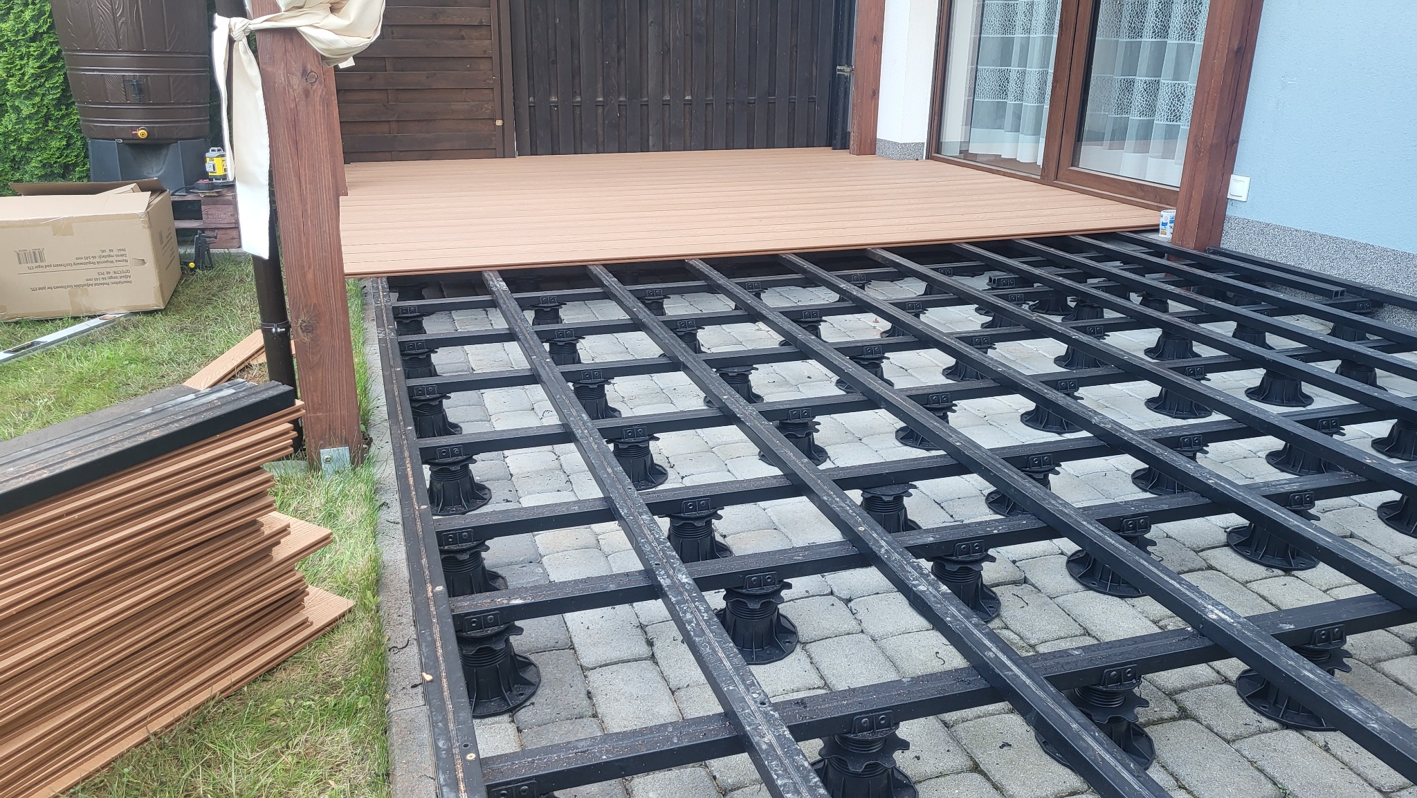 Montaż podkonstrukcji tarasu na regulowanych wspornikach. Częściowo ułożone deski, widoczne legary i kostka brukowa. Na trawie stos desek i karton.