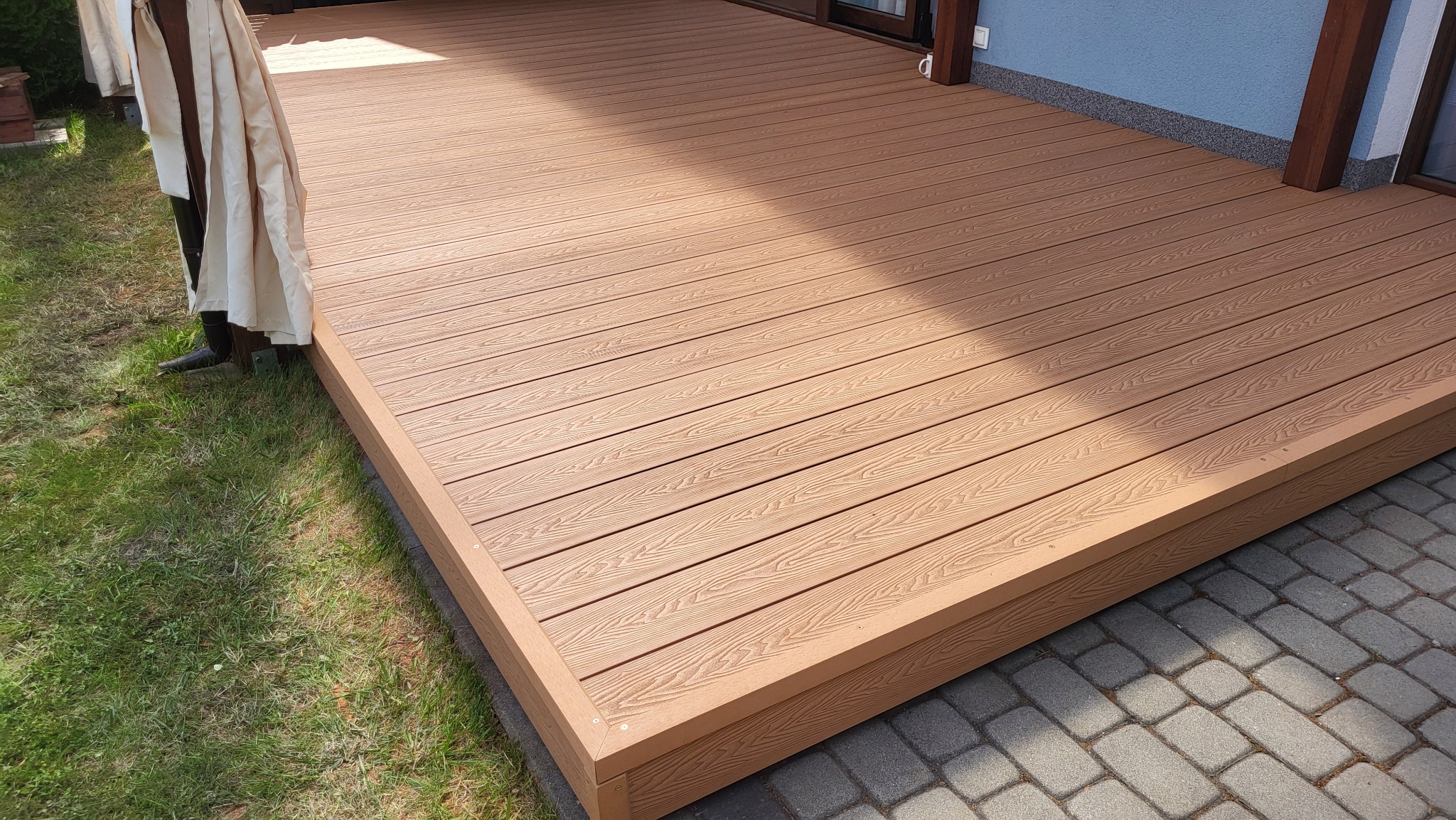 Jasnobrązowy taras z desek kompozytowych, z widoczną strukturą drewna, przylegający do domu z niebieską elewacją i fragmentem trawnika. Ujęcie z niskiej perspektywy.