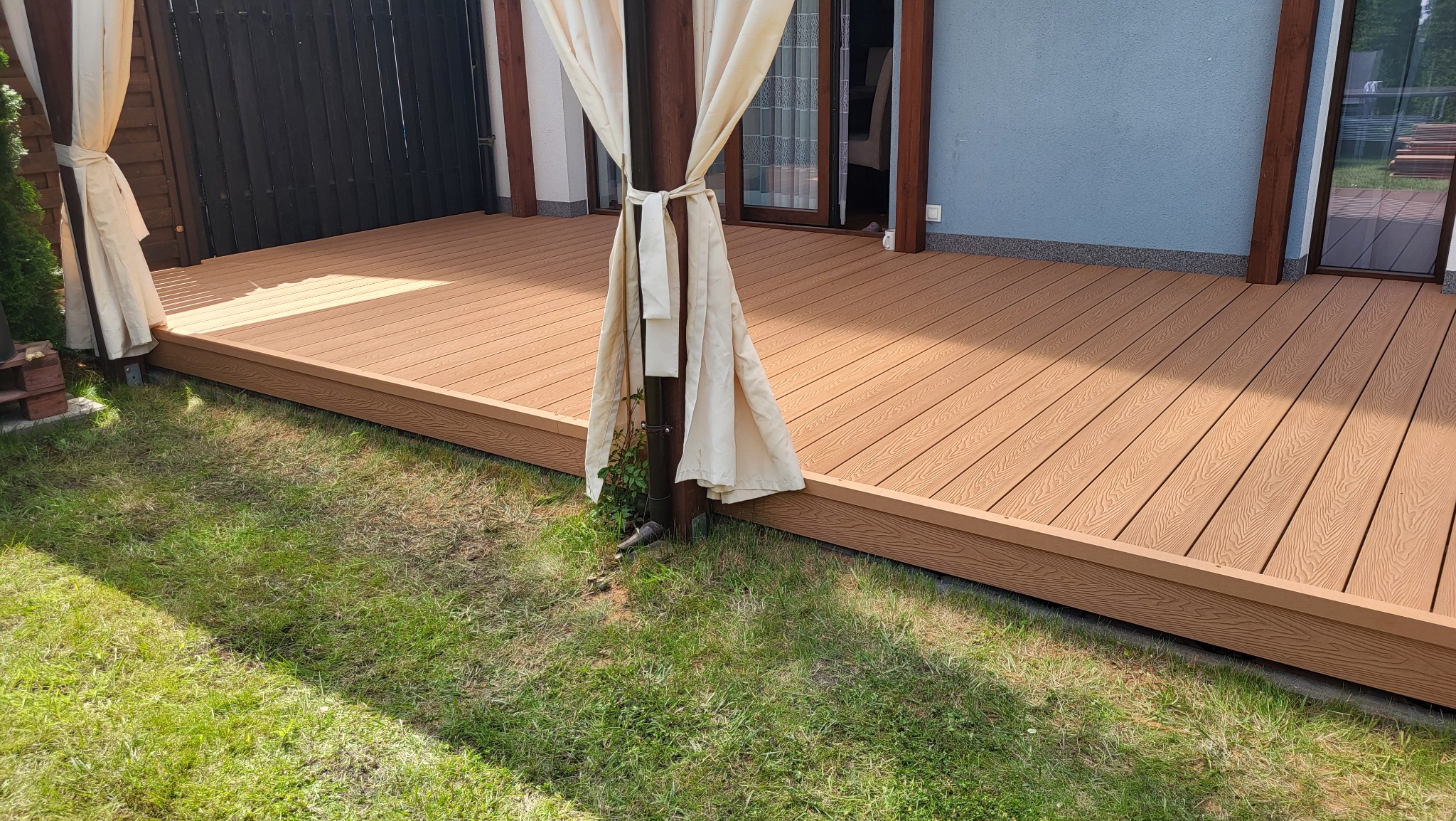 Jasnobrązowy taras z desek kompozytowych, zadaszony pergolą z beżowymi zasłonami, otoczony zielonym trawnikiem. Widok na fragment domu z niebieską elewacją.