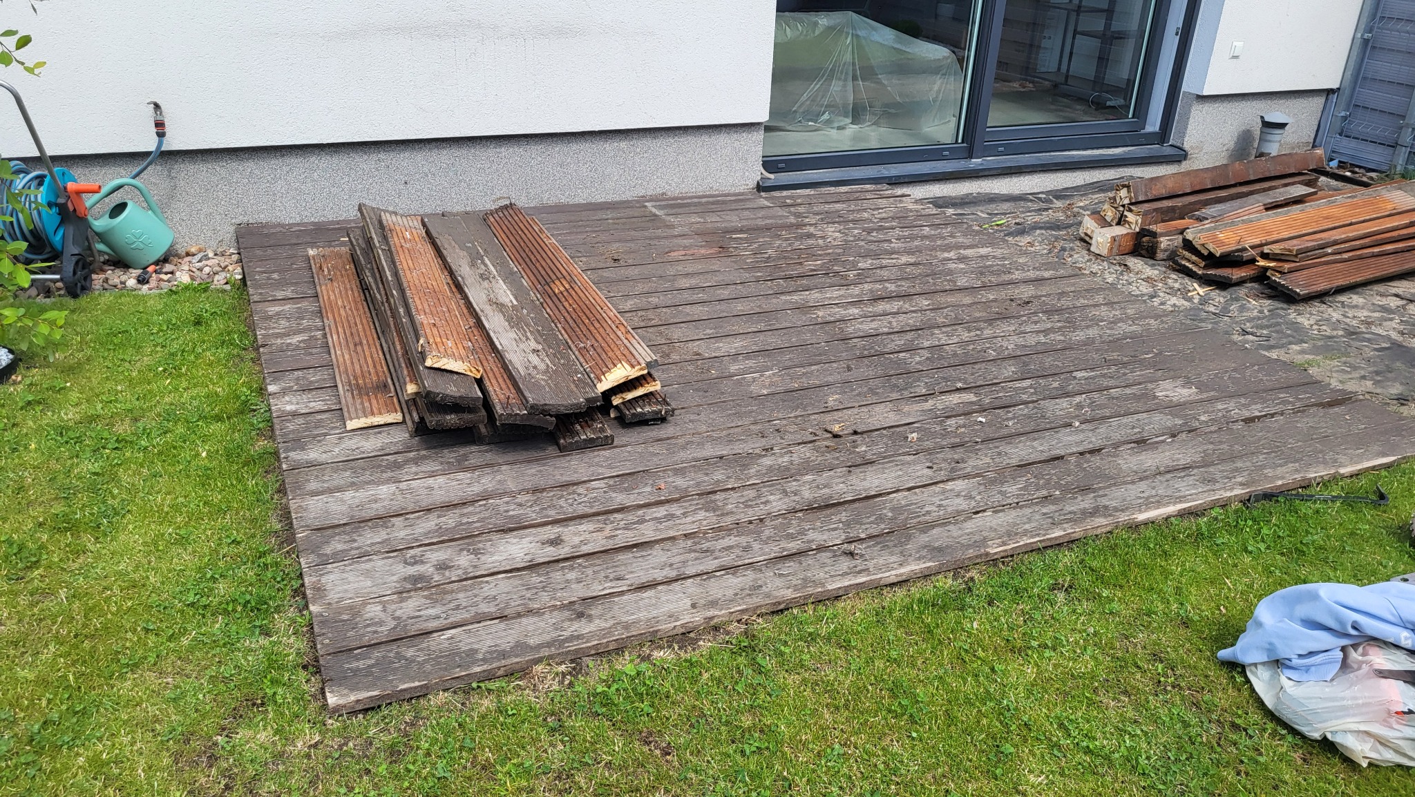 Stary, częściowo zdemontowany taras z desek przy domu, z widocznymi deskami do wymiany i narzędziami ogrodowymi w tle. Widok z poziomu trawnika.