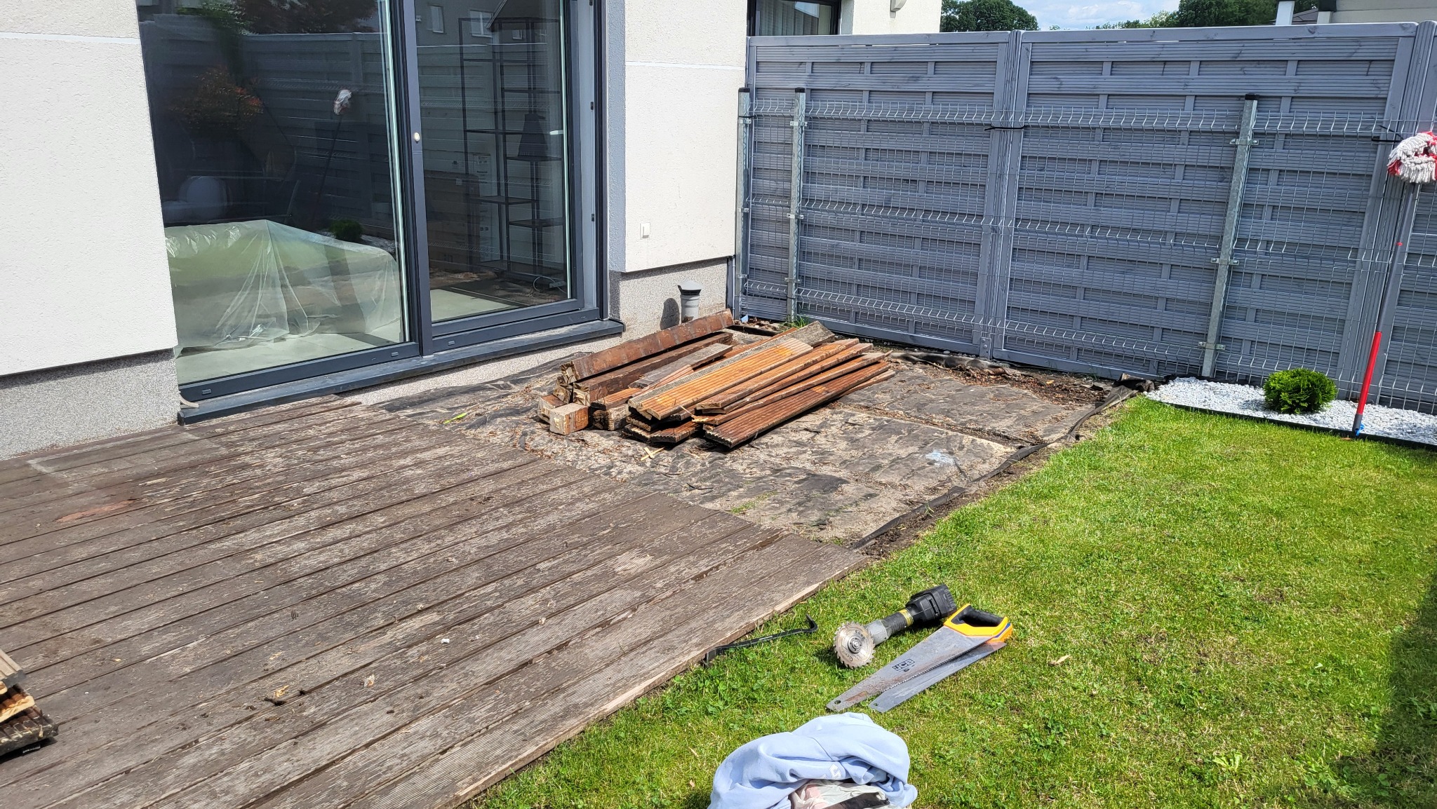 Demontaż starego tarasu drewnianego: deski, narzędzia (szlifierka kątowa, piła ręczna) na trawie obok częściowo zdemontowanego tarasu przy domu z szarym ogrodzeniem.