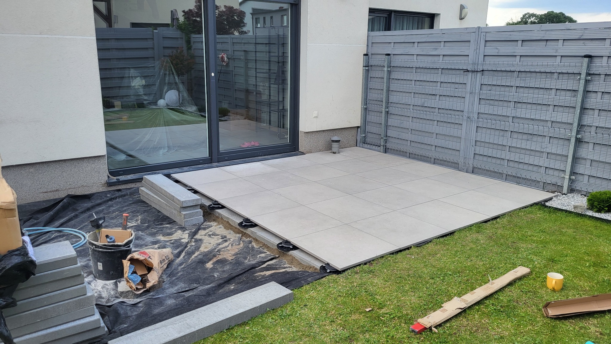 Montaż tarasu z płyt na regulowanych wspornikach. Częściowo ułożona powierzchnia, widoczne narzędzia i materiały budowlane. Szara kolorystyka, nowoczesny design.