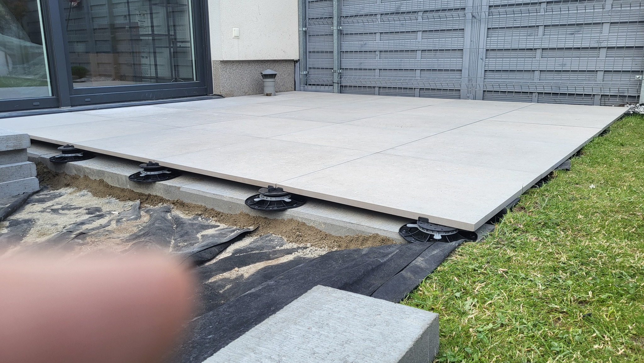 Montaż tarasu z płyt na regulowanych wspornikach. Widoczne warstwy podłoża i trawa. Płyty w jasnym odcieniu szarości, tworzące równą powierzchnię.