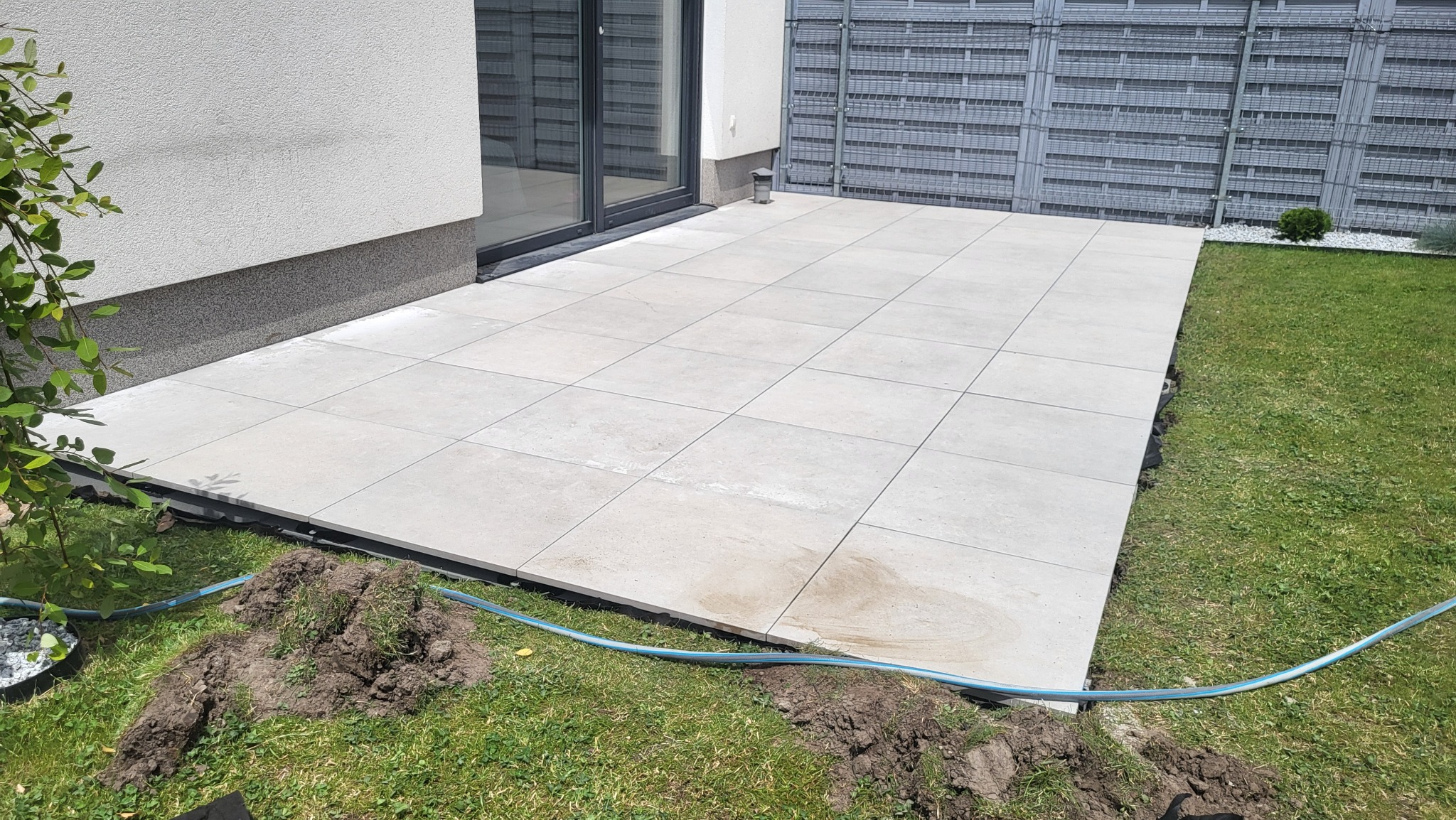 Jasny taras z płyt ułożonych na kwadratowo, przylegający do budynku z szarą elewacją, obok trawnik i szara ażurowa ścianka oddzielająca przestrzeń.