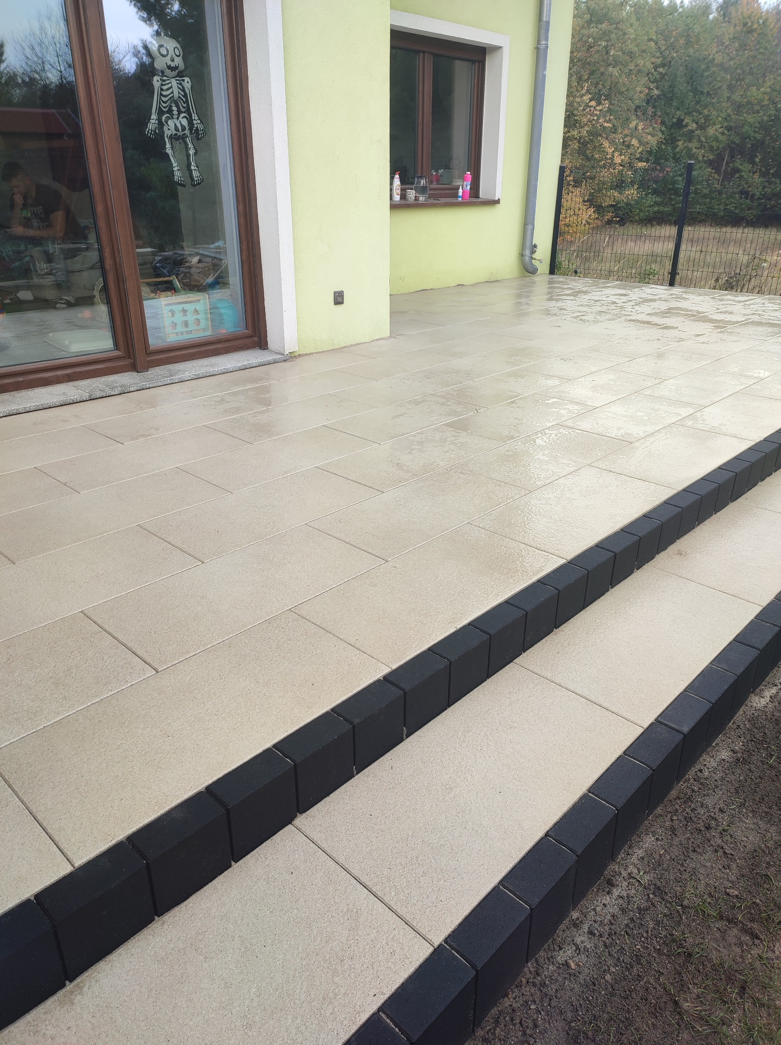 Jasna kostka brukowa na tarasie z czarnymi obrzeżami, widoczne schody, w tle okno domu z dekoracją Halloween