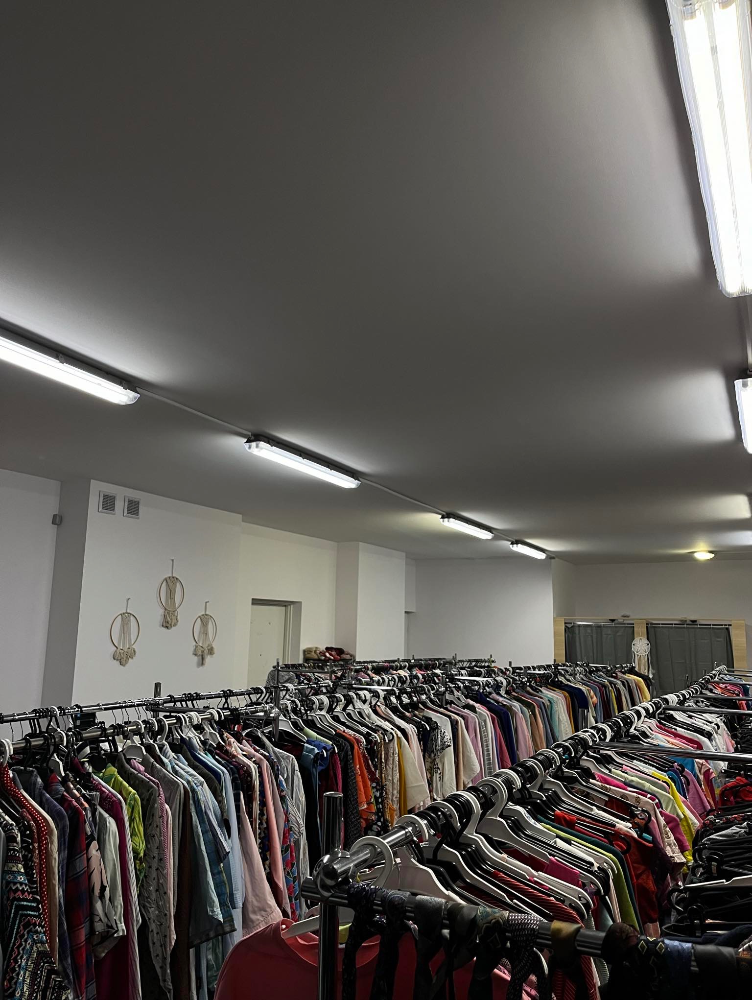 Wnętrze sklepu z odzieżą używaną, rzędy ubrań na wieszakach, widoczne dekoracje makramowe na ścianie i oświetlenie sufitowe.