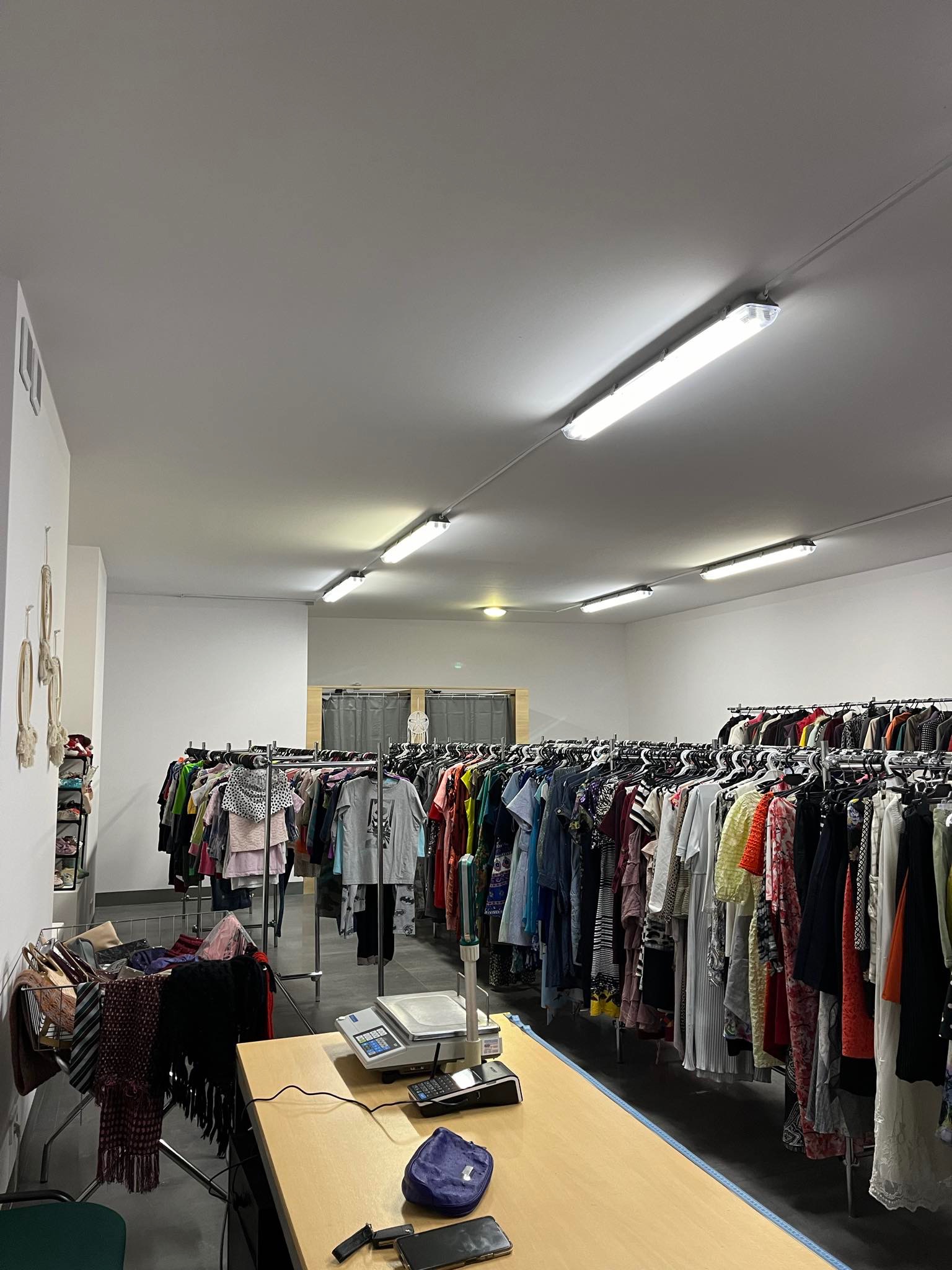 Wnętrze sklepu z odzieżą, rzędy ubrań na wieszakach, kasa fiskalna na ladzie, w tle okno z zasłoną.