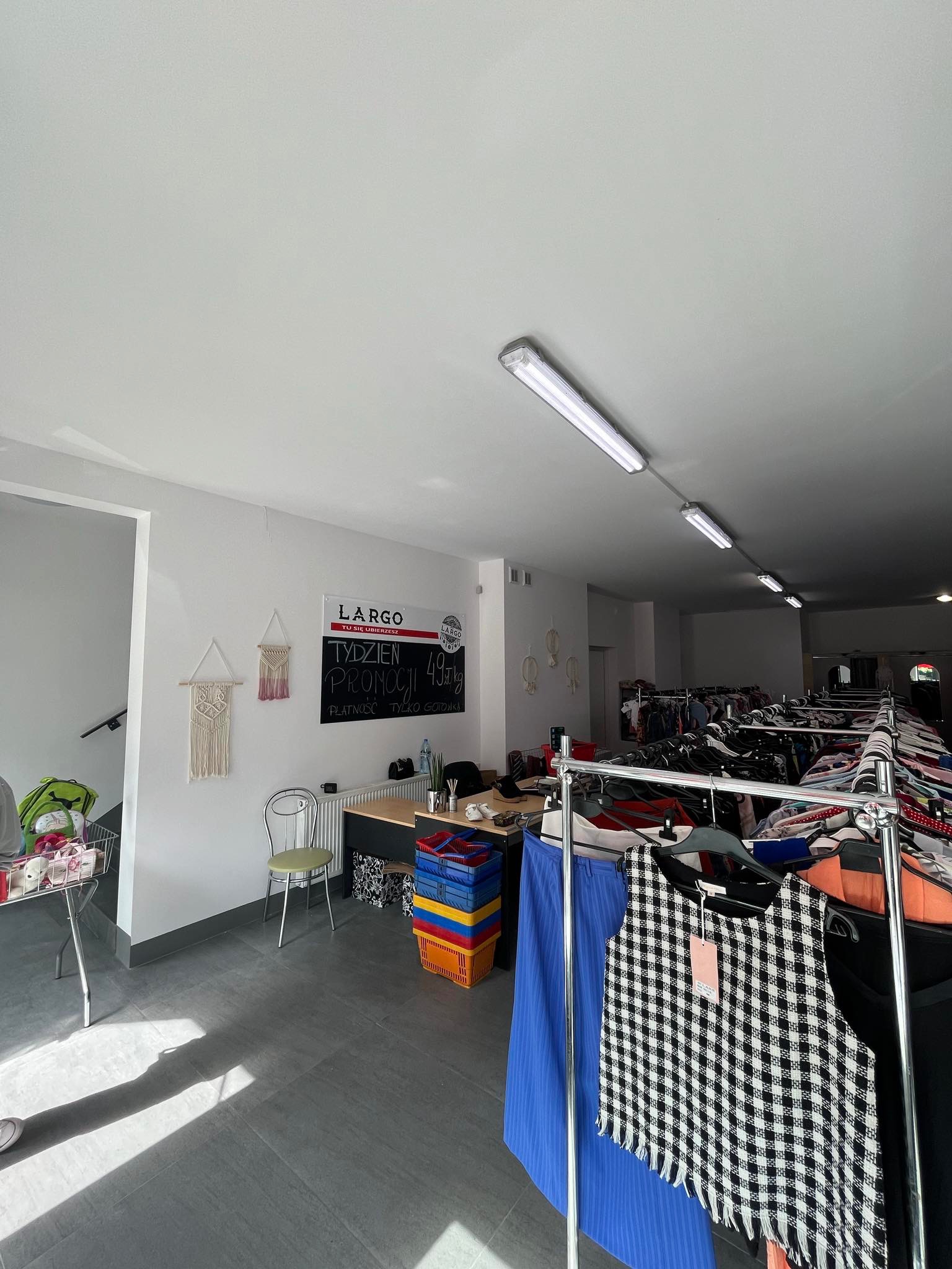 Wnętrze sklepu z odzieżą używaną, wieszaki z ubraniami, tablica informująca o promocji 'Tydzień Promocji', płatność tylko gotówką.