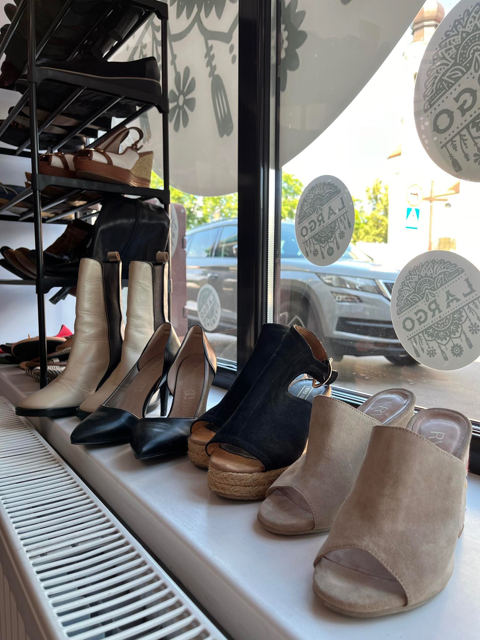Wystawa butów damskich w witrynie sklepowej, widoczne kozaki, szpilki, sandały na koturnie i klapki, na tle ulicy i dekoracyjnych naklejek na szybie.