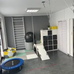 Sala rehabilitacyjna z drabinką, piłką, trampoliną, tablicą posturalną i elementami do ćwiczeń sensorycznych.