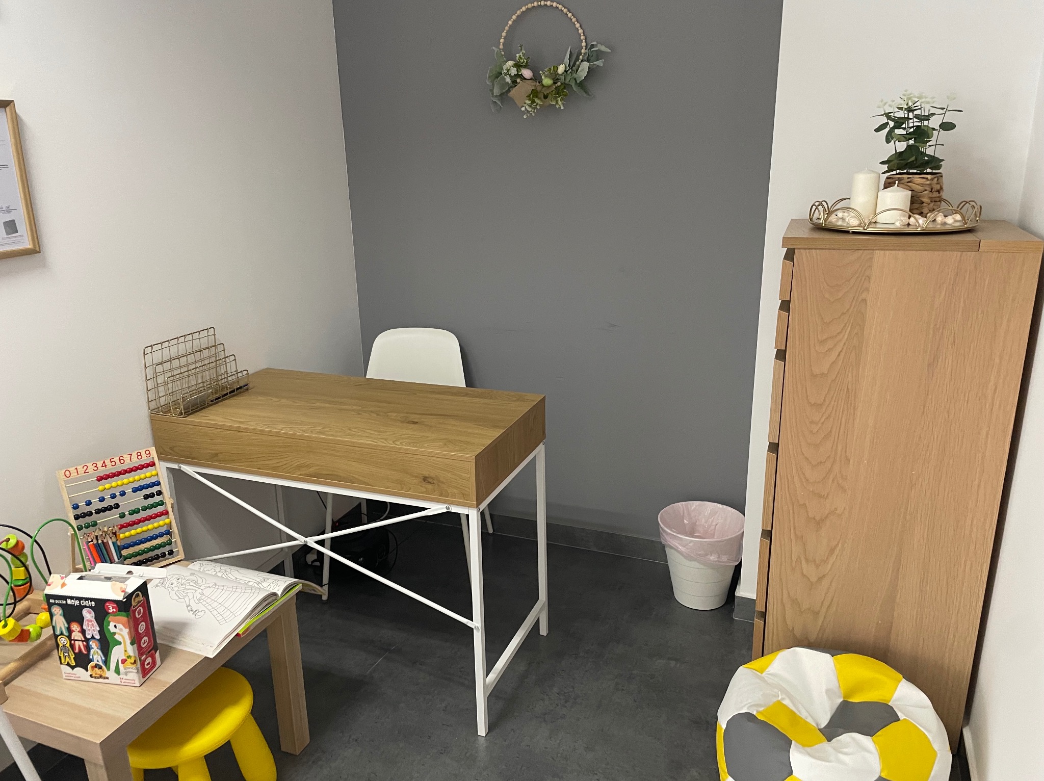 Jasne, minimalistyczne wnętrze gabinetu z drewnianym biurkiem na białych nogach, dziecięcym stolikiem z zabawkami edukacyjnymi i kolorowanką, dekoracjami roślinnymi na ścianie i szafce, oraz pufą...