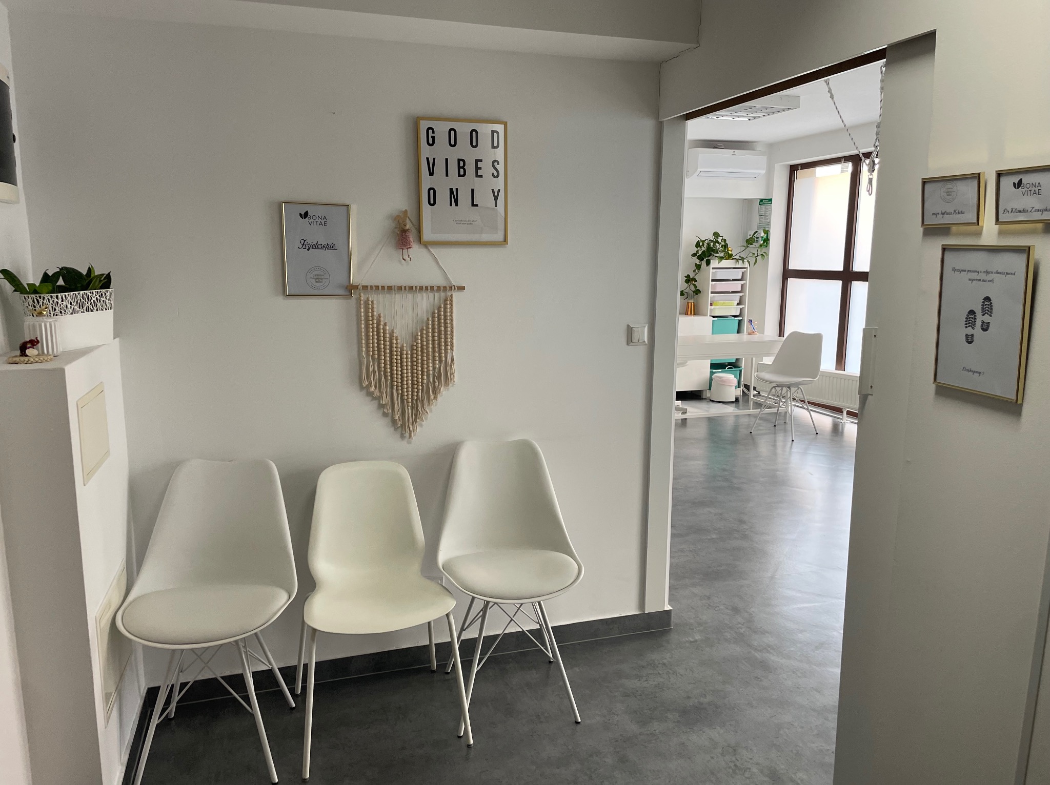 Poczekalnia w gabinecie z trzema białymi krzesłami, na ścianie dyplomy i plakat z napisem 'Good vibes only', widok na jasne pomieszczenie zabiegowe z białymi meblami.