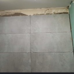 Prace Remontowo - Budowlane - Marcin Wójcik - Ściana w trakcie układania szarych płytek ceramicznych z krzyżykami dystansowymi, widoczne ślady kleju i niezabezpieczona płyta kartonowo-gipsowa.