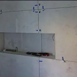 Ściana z szarych płytek ceramicznych w trakcie montażu, z widocznymi niebieskimi klipsami poziomującymi i wnęką z narzędziami.