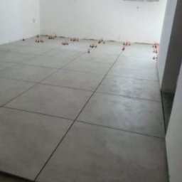 Prace Remontowo - Budowlane - Marcin Wójcik - Ułożone, szare płytki podłogowe w trakcie montażu, z widocznymi pomarańczowymi klipsami poziomującymi, w surowym wnętrzu z białymi ścianami i oknem.