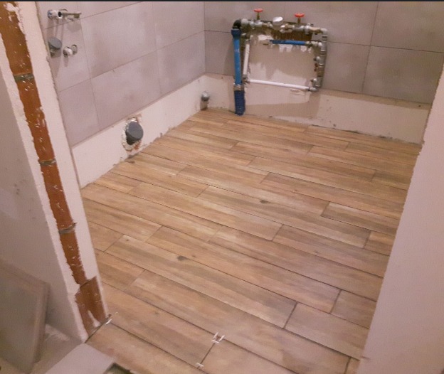 Ułożone drewnopodobne płytki podłogowe w łazience w trakcie remontu, widoczne instalacje wodne i kanalizacyjne, oraz fragment nie wykończonego ościeża drzwi.