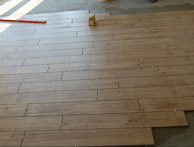 Układanie podłogi z płytek imitujących drewno, z zastosowaniem krzyżyków dystansowych, widoczny poziomica i gąbka.