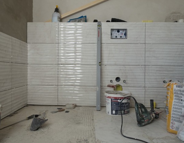 Ściana w trakcie wykańczania z ułożonymi płytkami o falistej strukturze, widoczne otwory na instalacje, poziomica, wiadro z klejem i narzędzia na podłodze.