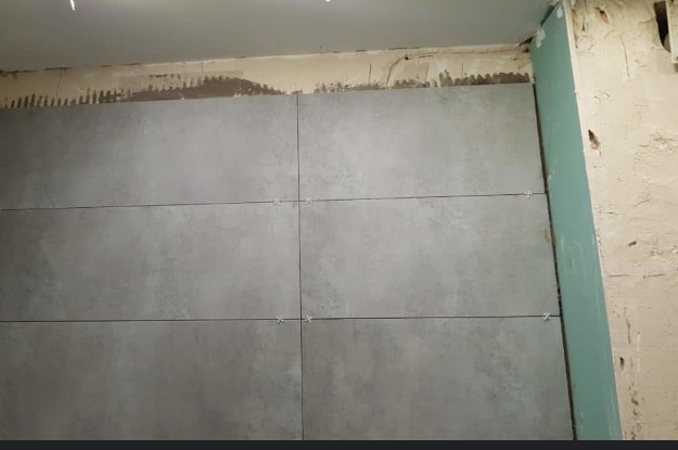 Ściana w trakcie układania szarych płytek ceramicznych z krzyżykami dystansowymi, widoczne ślady kleju i niezabezpieczona płyta kartonowo-gipsowa.