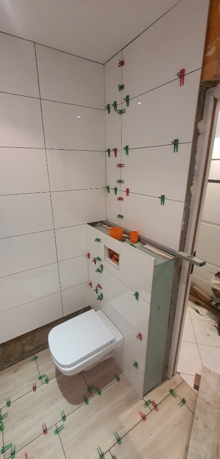 Białe płytki ścienne z zielonymi i czerwonymi klipsami poziomującymi podczas układania w łazience, widoczny stelaż podtynkowy WC, płytki podłogowe w trakcie montażu.