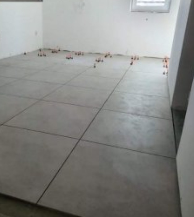 Ułożone, szare płytki podłogowe w trakcie montażu, z widocznymi pomarańczowymi klipsami poziomującymi, w surowym wnętrzu z białymi ścianami i oknem.