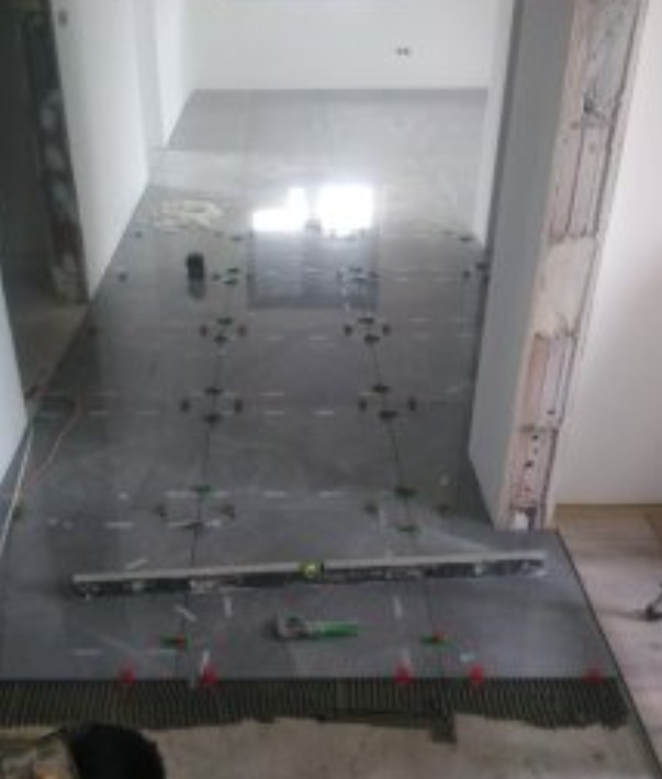 Układanie ciemnoszarych płytek podłogowych w korytarzu, widoczne poziomica, krzyżyki dystansowe i fragment grzebienia do rozprowadzania kleju.