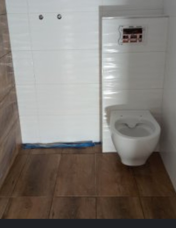 Montaż podwieszanej toalety z białą ceramiką na brązowej podłodze imitującej drewno, białe płytki na ścianach, widoczne elementy instalacji.