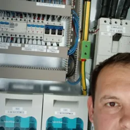 Elektryk częściowo zasłania rozdzielnicę elektryczną z bezpiecznikami, przewodami i dwoma licznikami energii umieszczonymi w obudowach z tworzywa sztucznego.