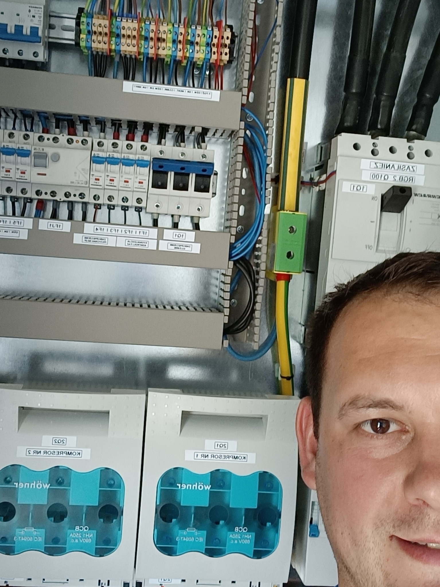 Elektryk częściowo zasłania rozdzielnicę elektryczną z bezpiecznikami, przewodami i dwoma licznikami energii umieszczonymi w obudowach z tworzywa sztucznego.