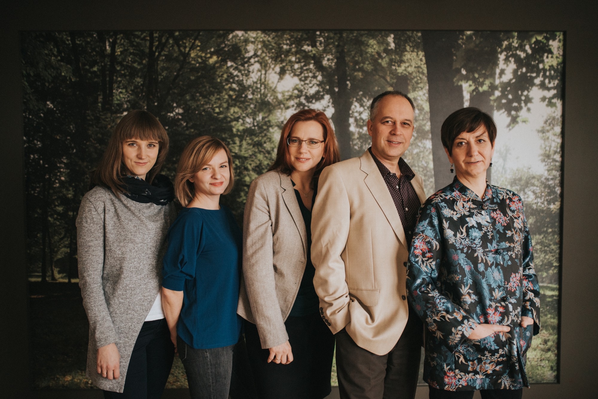 Grupa pięciu profesjonalistów w różnym wieku, stojących ramię w ramię na tle grafiki przedstawiającej las, z delikatnymi uśmiechami i profesjonalnym wyglądem.