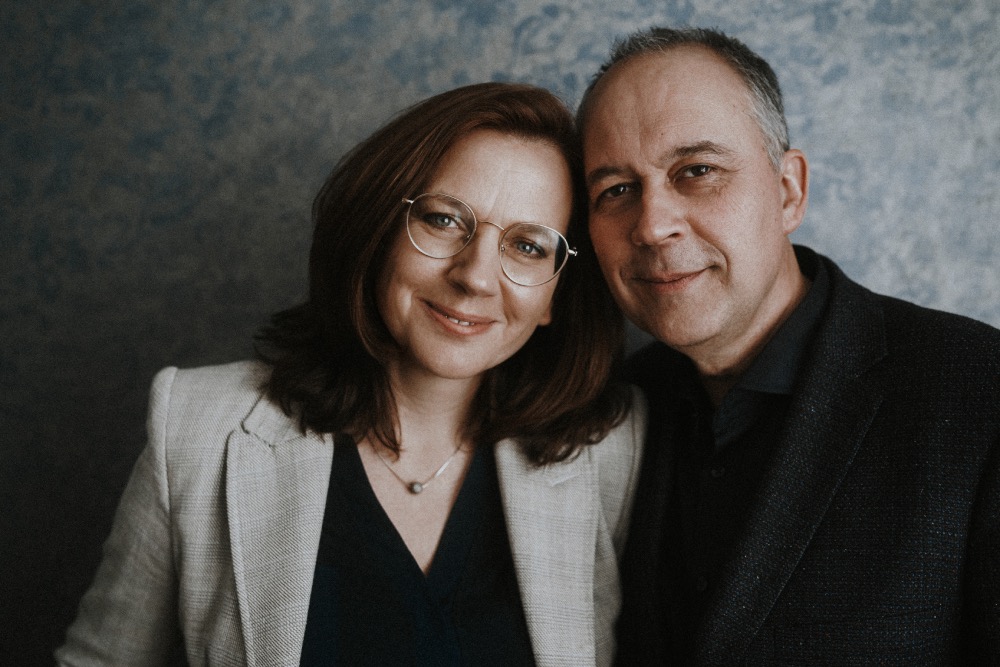 Portret uśmiechniętej kobiety w okularach i beżowej marynarce, stojącej obok mężczyzny w ciemnej marynarce na tle teksturowanej ściany.