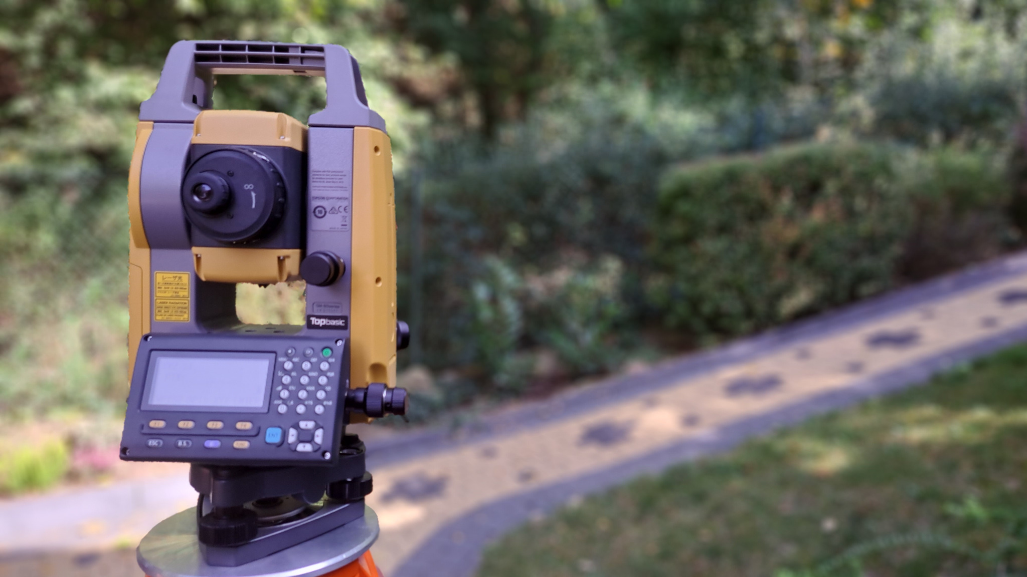 Żółto-szary teodolit Topbasic ustawiony na statywie, w tle rozmyta ścieżka i zieleń.