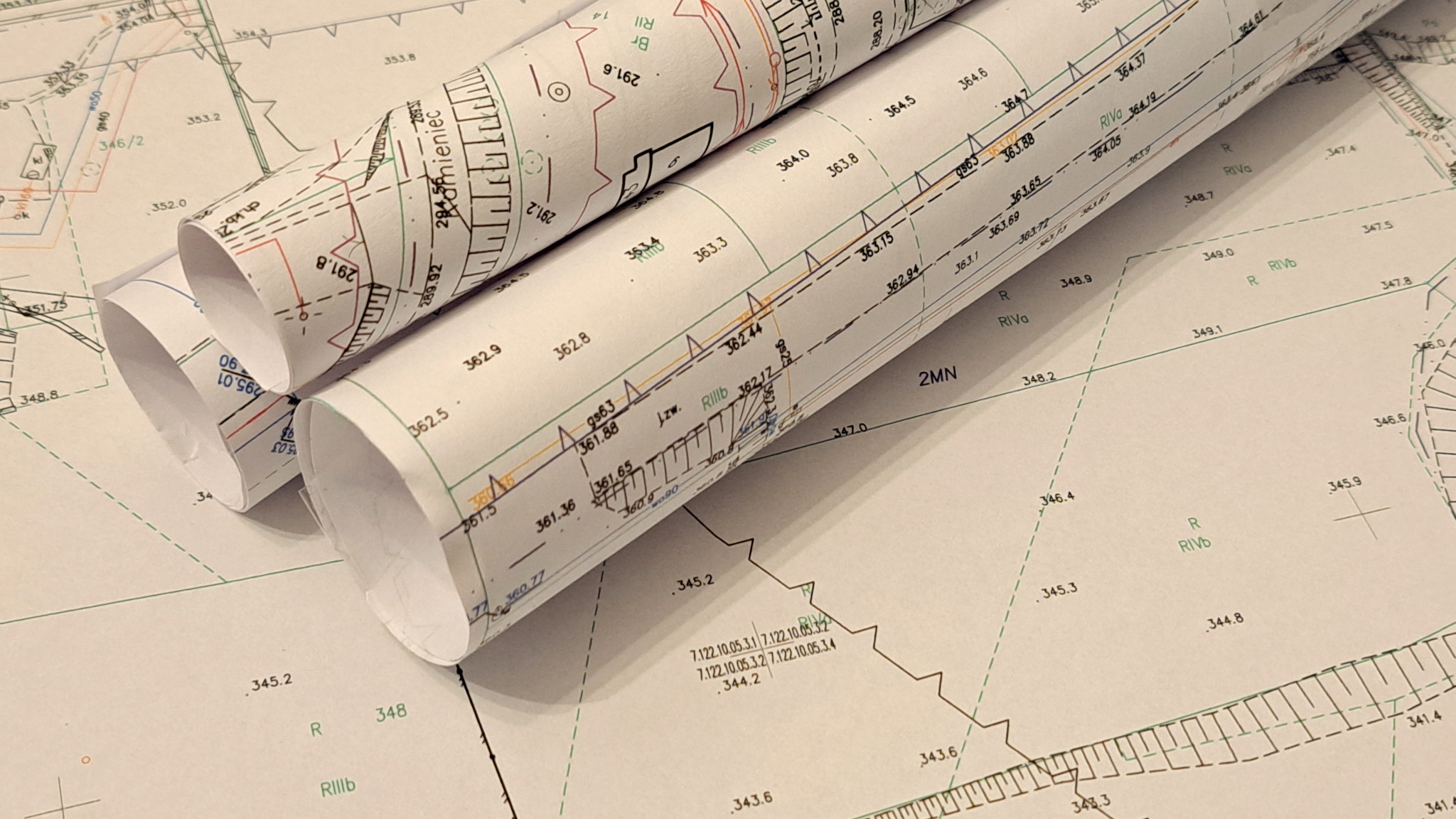 Zrolowane plany geodezyjne z naniesionymi numerami działek i wysokościami terenu, leżące na rozłożonym planie. Widoczne szczegółowe oznaczenia i linie pomiarowe.