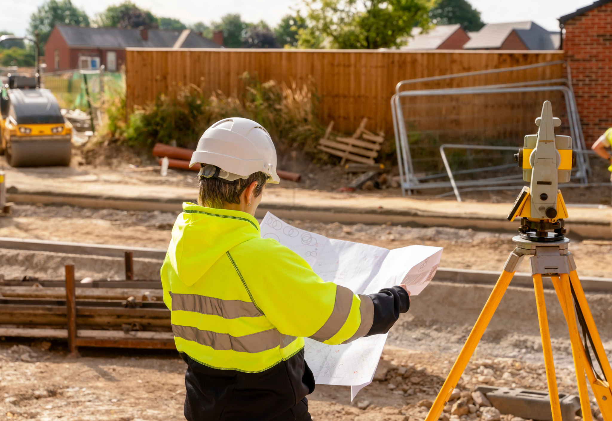 Geodeta w żółtej kamizelce i kasku analizuje plan na budowie, z niwelatorem na statywie w tle i walcem drogowym w oddali.