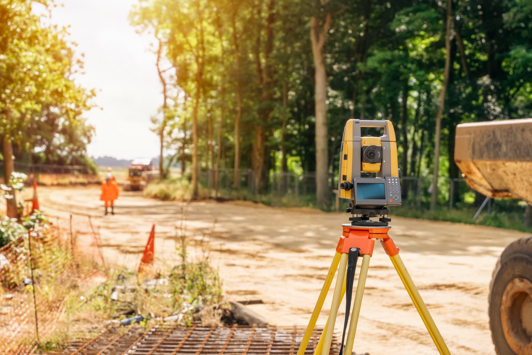 Żółty teodolit na trójnogu, ustawiony na placu budowy z widocznym operatorem w pomarańczowym kombinezonie i ciężkim sprzętem w tle, oświetlony promieniami słonecznymi.