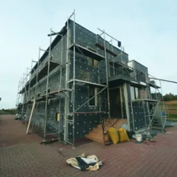 Dom w trakcie ocieplania elewacji, widoczne rusztowania, płyty izolacyjne z białymi kołkami, worki z materiałami budowlanymi i częściowo wykończona fasada przy wejściu.