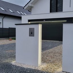 VADYM HAL DOBRO W DZIAŁANIU - Nowoczesna elewacja domu jednorodzinnego z białym tynkiem, oświetlonymi słupkami ogrodzeniowymi i szarą bramą garażową.